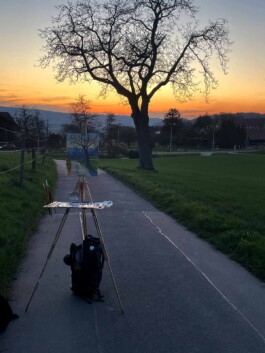 Die Sonne ist gerade untergegangen, vor dem leuchtenden Orange des Himmels steht ein Baum ohne Blätter. Das Gemälde davon sieht man klein auf einer Feldstaffelei. 